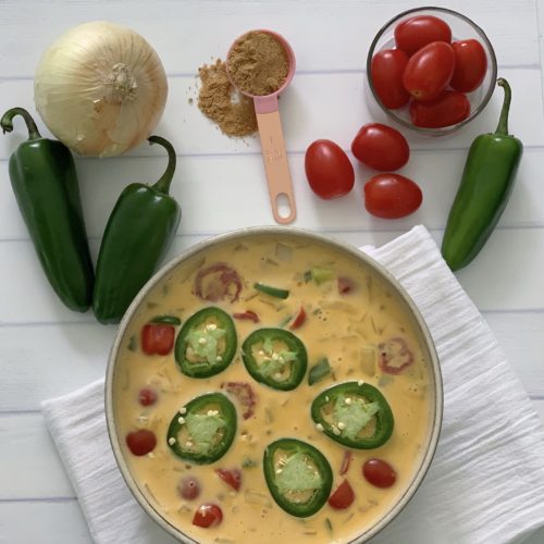 An Authentic Easy Queso Dip Recipe featured by top US food blog, Food Diary of a City Girl: image of a bowl with queso dip surrounded by onion, three peppers, and cherry tomatoes.