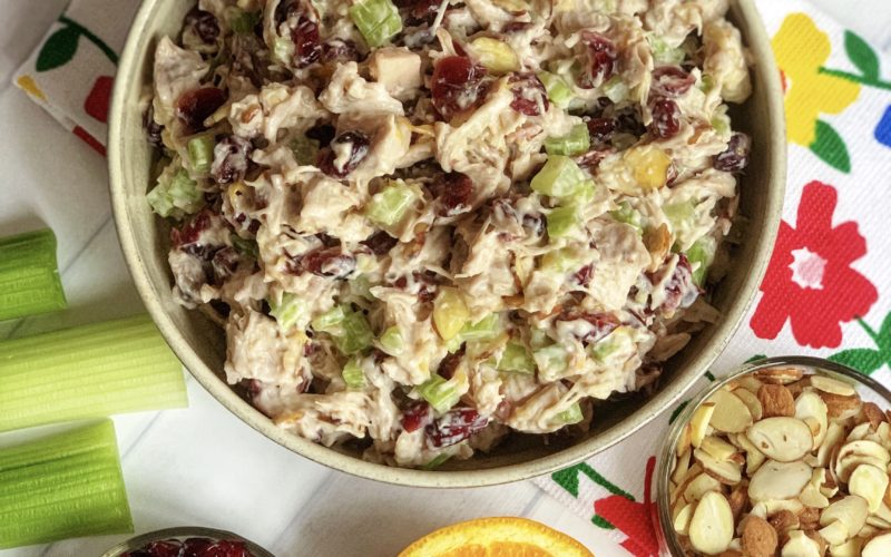 Quick Summer Salad Idea: Cranberry Almond Chicken Salad Recipe featured by top US food blog, Food Diary of a City Girl: image of a pottery bowl with Cranberry Almond Chicken Salad.  The bowl sits on top a floral kitchen towel.  Next to the Cranberry-Almond Chicken Salad are three celery stalks, half of an orange, a clear bowl of almonds, and a clear bowl of dried cranberries.