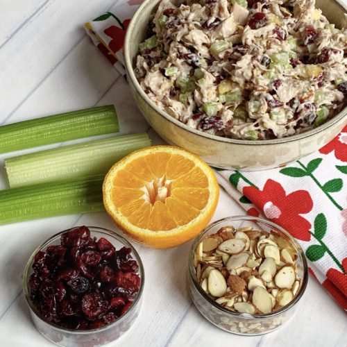 Quick Summer Salad Idea: Cranberry Almond Chicken Salad Recipe featured by top US food blog, Food Diary of a City Girl: image of a pottery bowl with Cranberry Almond Chicken Salad.  The bowl sits on top a floral kitchen towel.  Next to the Cranberry-Almond Chicken Salad are three celery stalks, half of an orange, a clear bowl of almonds, and a clear bowl of dried cranberries.