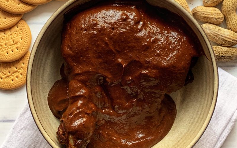 Quick and Easy Mole Recipe featured by top US food blog, Food Diary of a City Girl: image of a pottery bowl with chicken and mole sauce sitting atop a white kitchen linen towel and surrounded by small clear glass container with Cumin Seeds, Maria Cookies, and Peanuts.