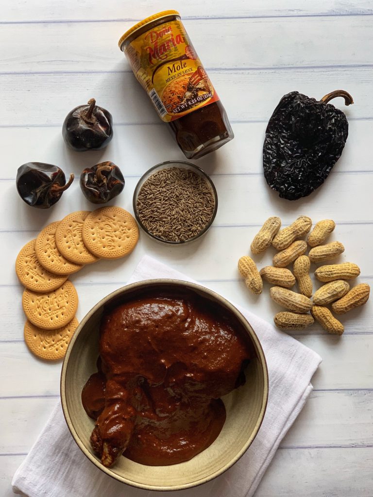 Quick and Easy Mole Recipe featured by top US food blog, Food Diary of a City Girl: image of a pottery bowl with chicken and mole sauce sitting atop a white kitchen linen towel and surrounded by small clear glass container with Cumin Seeds, Maria Cookies, and Peanuts.