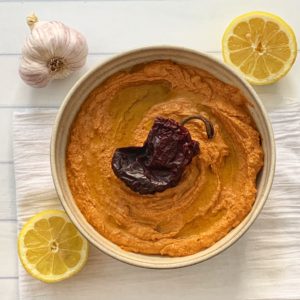 Spicy Hummus Recipe: Delicious Ancho Chili Hummus featured by top US food blog, Food Diary of a City Girl: image of pottery bowl with ancho chili hummus topped with an ancho chili, surrounded by a two lemon halves, and one garlic bulb.