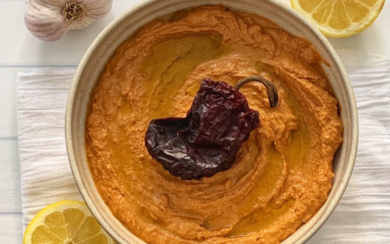 Spicy Hummus Recipe: Delicious Ancho Chili Hummus featured by top US food blog, Food Diary of a City Girl: image of pottery bowl with ancho chili hummus topped with an ancho chili, surrounded by a two lemon halves, and one garlic bulb.