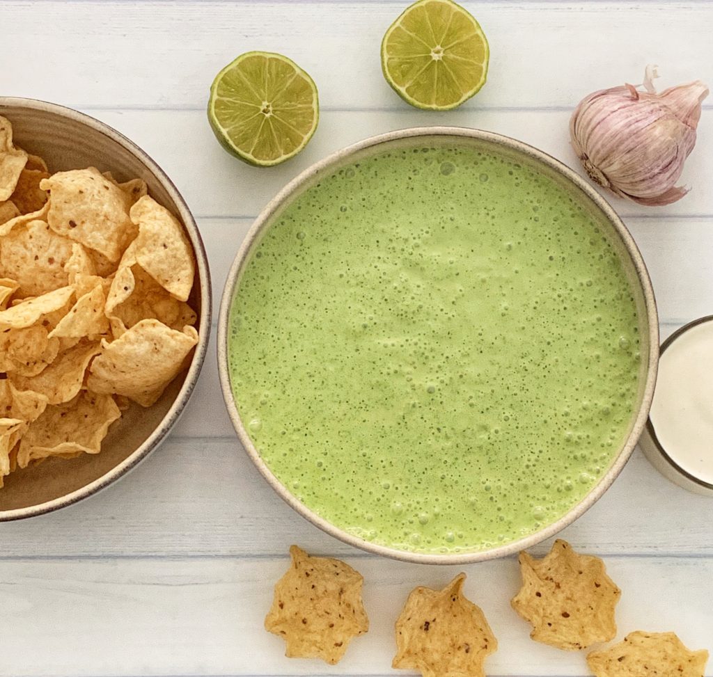 Poblano Pepper Crema: The Ultimate Poblano Cheese Dip Recipe featured by top US food blog, Food Diary of a City Girl: image of a bowl with poblano pepper crema surrounded by garlic, two lime halves, a small glass container with sour cream, and a bowl of corn tortilla chips.