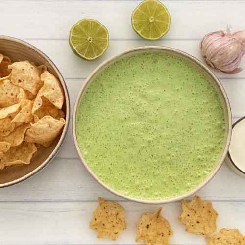 Poblano Pepper Crema: The Ultimate Poblano Cheese Dip Recipe featured by top US food blog, Food Diary of a City Girl: image of a bowl with poblano pepper crema surrounded by garlic, two lime halves, a small glass container with sour cream, and a bowl of corn tortilla chips.