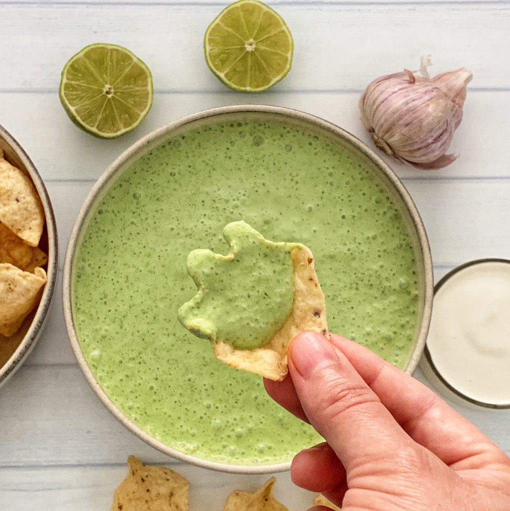 Poblano Pepper Crema: The Ultimate Poblano Cheese Dip Recipe featured by top US food blog, Food Diary of a City Girl: image of a bowl with poblano pepper crema surrounded by garlic, two lime halves, a small glass container with sour cream, and a hand with one chip dipping in the Poblano Pepper Crema.