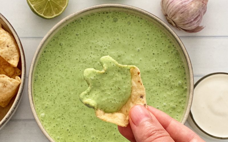 Poblano Pepper Crema: The Ultimate Poblano Cheese Dip Recipe featured by top US food blog, Food Diary of a City Girl: image of a bowl with poblano pepper crema surrounded by garlic, two lime halves, a small glass container with sour cream, and a hand with one chip dipping in the Poblano Pepper Crema.