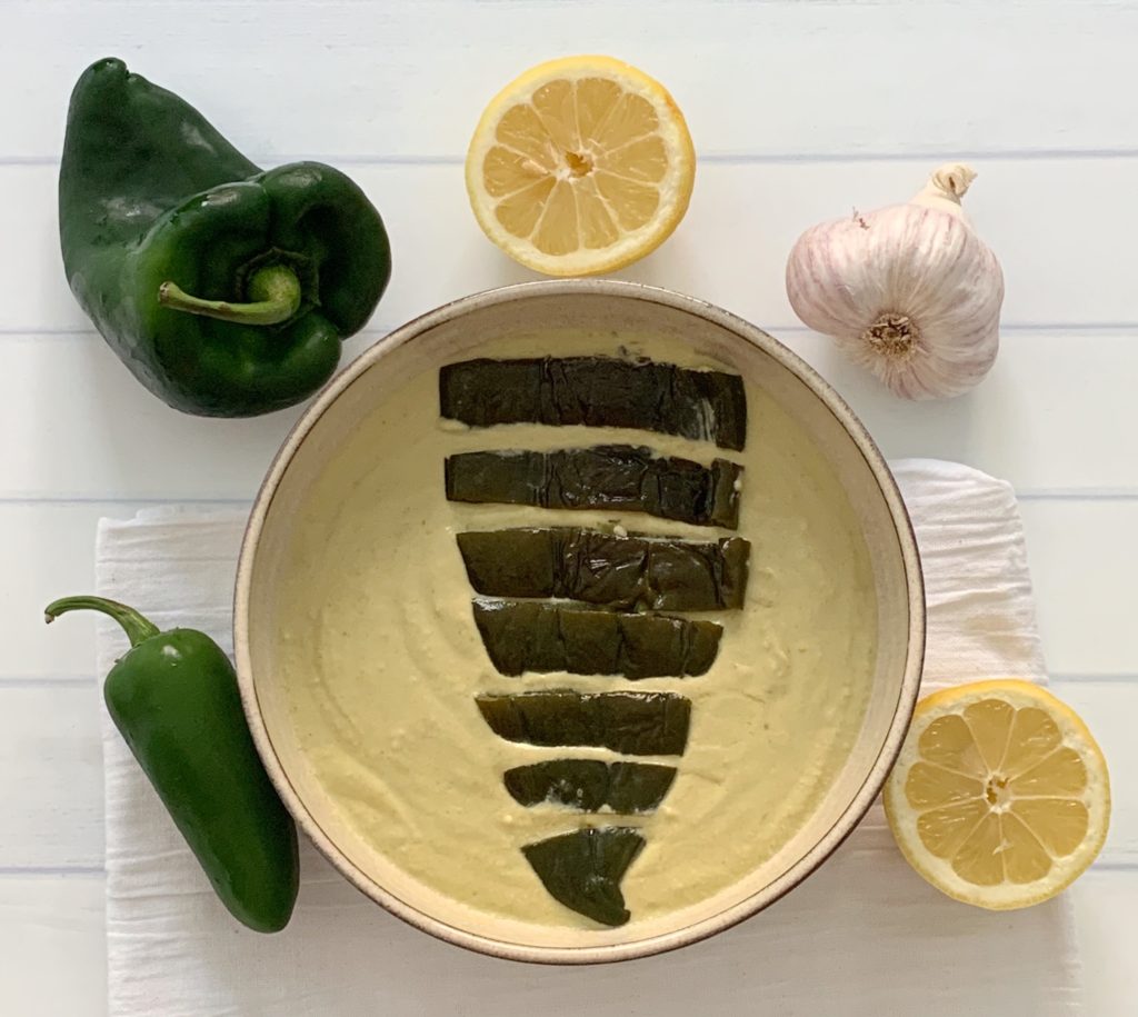 Award Winning Roasted Poblano Hummus Recipe featured by top US food blog, Food Diary of a City Girl: image of pottery bowl with roasted poblano hummus topped with a roasted poblano, surrounded by a poblano pepper, a jalapeño pepper, two lemon halves, and one garlic bulb.