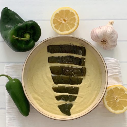 Award Winning Roasted Poblano Hummus Recipe featured by top US food blog, Food Diary of a City Girl: image of pottery bowl with roasted poblano hummus topped with a roasted poblano, surrounded by a poblano pepper, a jalapeño pepper, two lemon halves, and one garlic bulb.