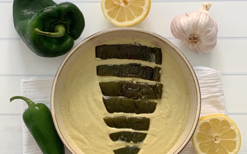 Award Winning Roasted Poblano Hummus Recipe featured by top US food blog, Food Diary of a City Girl: image of pottery bowl with roasted poblano hummus topped with a roasted poblano, surrounded by a poblano pepper, a jalapeño pepper, two lemon halves, and one garlic bulb.