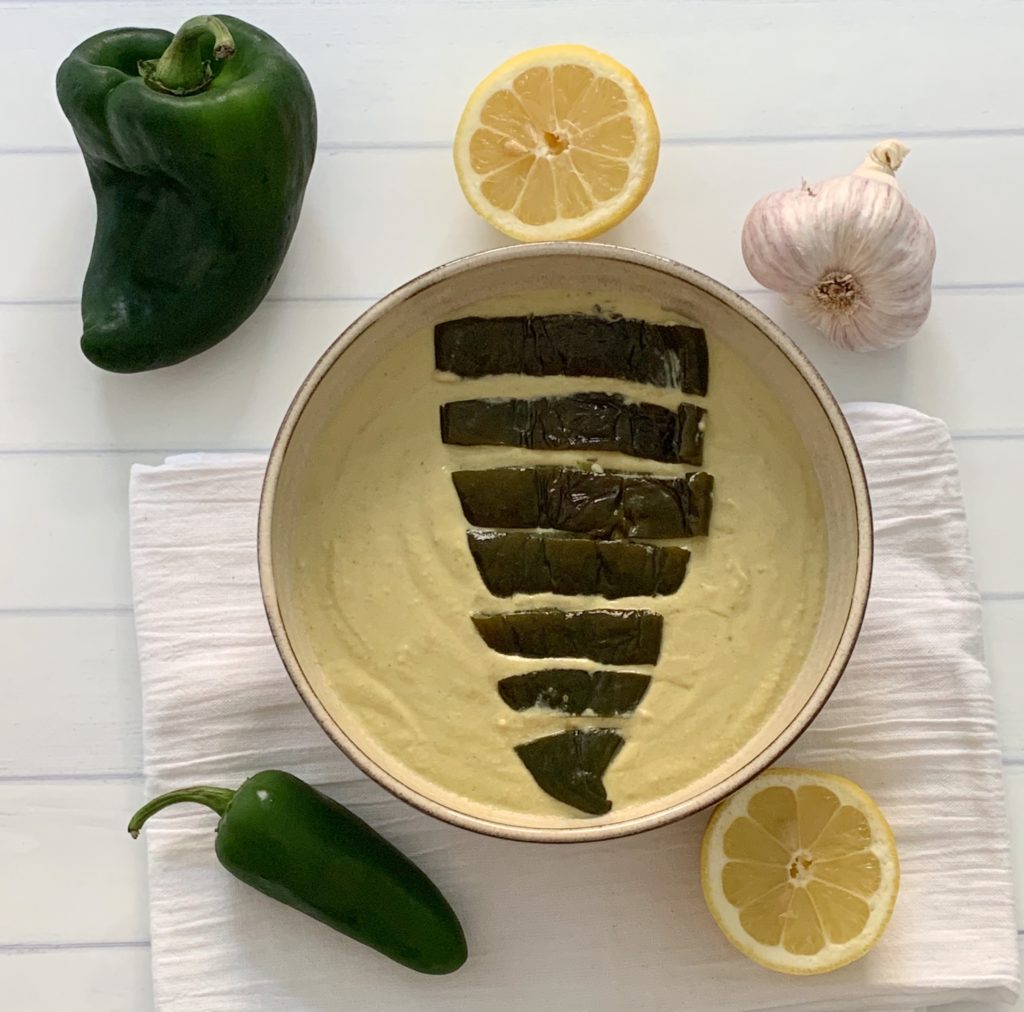 Award Winning Roasted Poblano Hummus Recipe featured by top US food blog, Food Diary of a City Girl: image of pottery bowl with roasted poblano hummus topped with a roasted poblano, surrounded by a poblano pepper, a jalapeño pepper, two lemon halves, and one garlic bulb.