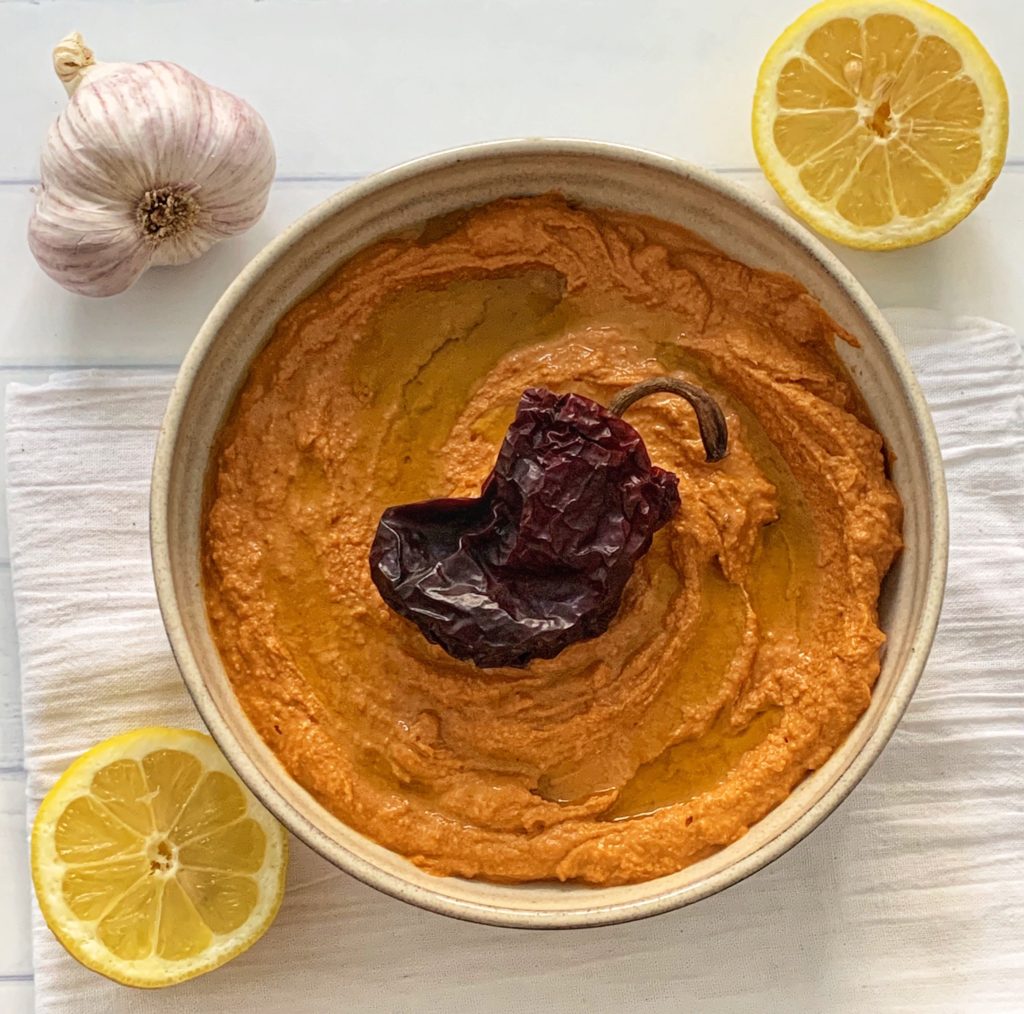 Spicy Hummus Recipe: Delicious Ancho Chili Hummus featured by top US food blog, Food Diary of a City Girl: image of pottery bowl with ancho chili hummus topped with an ancho chili, surrounded by a two lemon halves, and one garlic bulb.