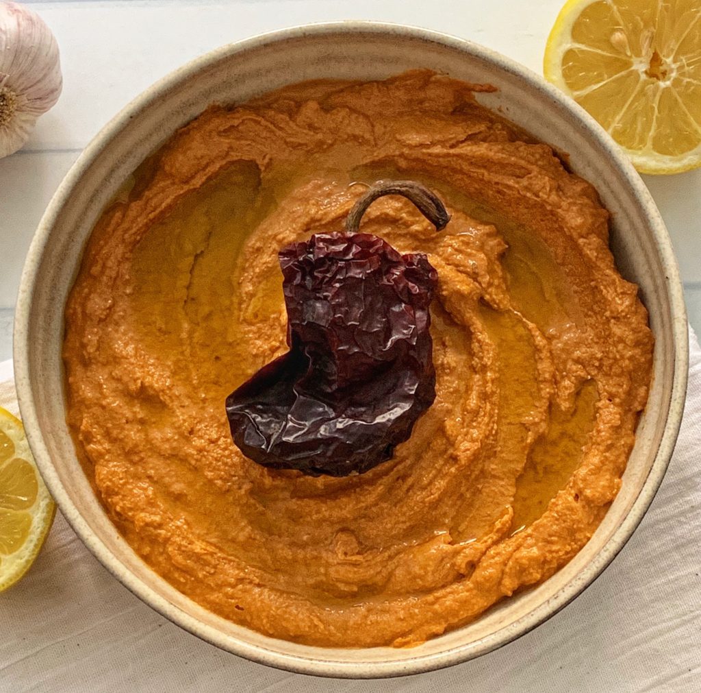 Spicy Hummus Recipe: Delicious Ancho Chili Hummus featured by top US food blog, Food Diary of a City Girl: image of pottery bowl with ancho chili hummus topped with an ancho chili, surrounded by a two lemon halves, and one garlic bulb.