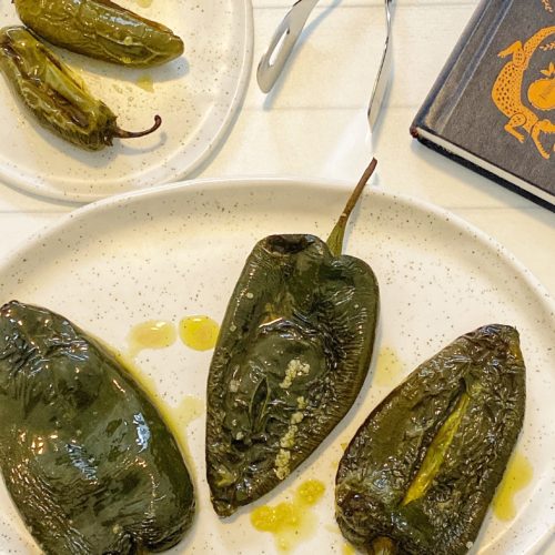 How to Roast Chili Peppers in the Oven: 5 Easy Steps featured by top US food blog, Food Diary of a City Girl: image of three roasted poblano peppers on a speckled pottery plate, three roasted jalapeño peppers on a small speckled pottery plate, silver tongs, and a blue book.