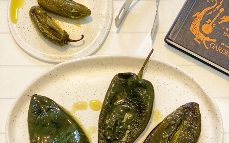 How to Roast Chili Peppers in the Oven: 5 Easy Steps featured by top US food blog, Food Diary of a City Girl: image of three roasted poblano peppers on a speckled pottery plate, three roasted jalapeño peppers on a small speckled pottery plate, silver tongs, and a blue book.