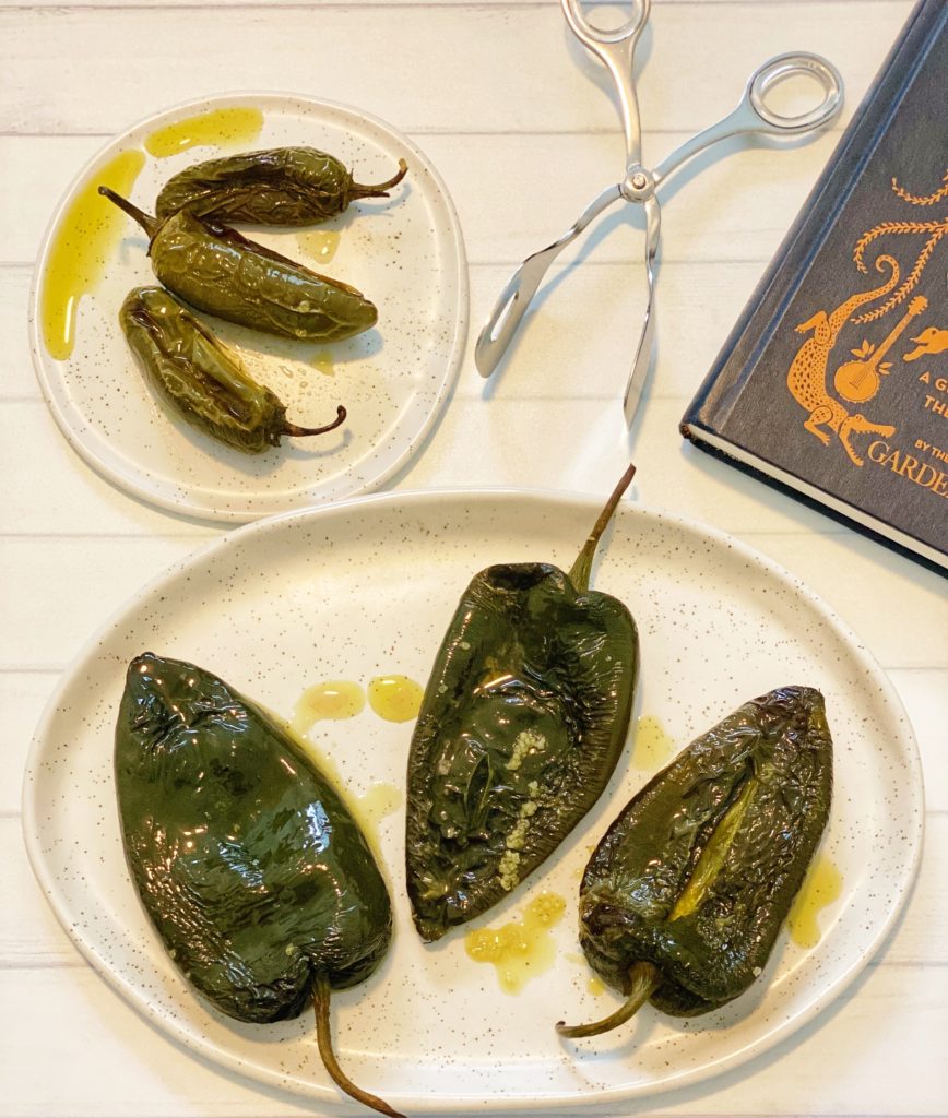 How to Roast Chili Peppers in the Oven: 5 Easy Steps featured by top US food blog, Food Diary of a City Girl: image of three roasted poblano peppers on a speckled pottery plate, three roasted jalapeño peppers on a small speckled pottery plate, silver tongs, and a blue book.