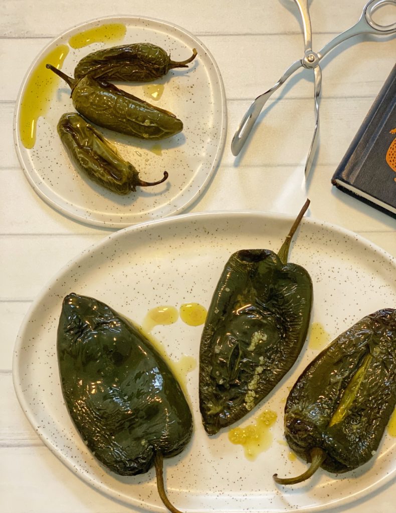 How to Roast Chili Peppers in the Oven: 5 Easy Steps featured by top US food blog, Food Diary of a City Girl: image of three roasted poblano peppers on a speckled pottery plate, three roasted jalapeño peppers on a small speckled pottery plate, silver tongs, and a blue book.